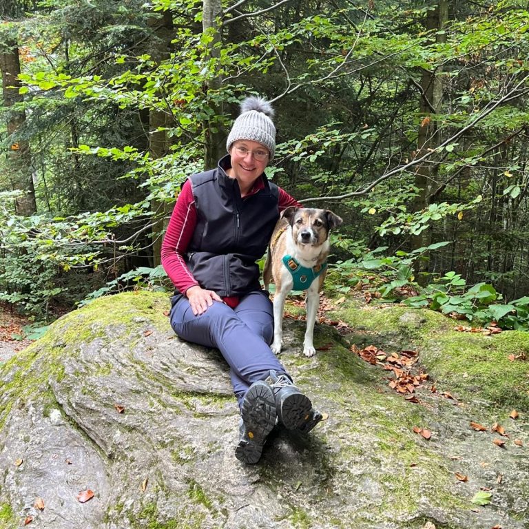 Lucy und Susanne Frau sitzt auf einem Felsen im Wald, neben ihr ein Hund. Herbstliche Farben um sie herum.
